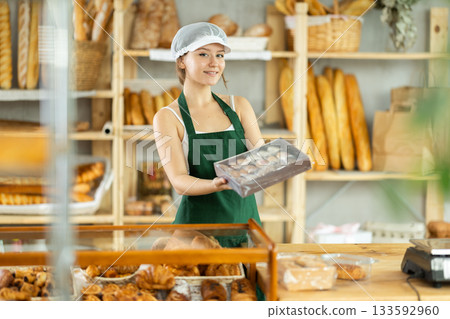 Nice young salesgirl in apron and cap offering cookies in bakery shop Nice young salesgirl in apron and cap offering cookies in bakery shop 133592960