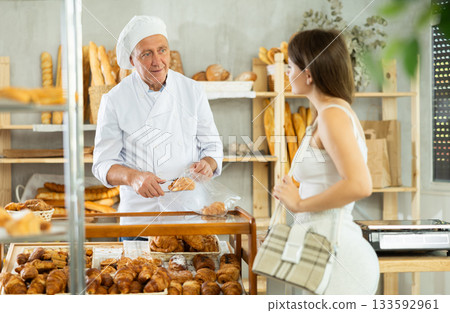 Girl selects and buys hot croissants at a bakery. Male salesperson places croissants in plastic bag Girl selects and buys hot croissants at a bakery. Male salesperson places croissants in plastic bag 133592961