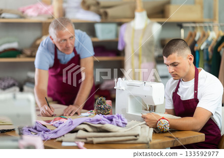 young tailor sews next to a mature employee of the atelier young tailor sews next to a mature employee of the atelier 133592977