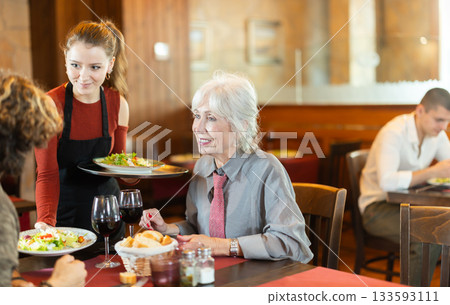 Young waitress bringing food to adult couple in restaurant Young waitress bringing food to adult couple in restaurant 133593111