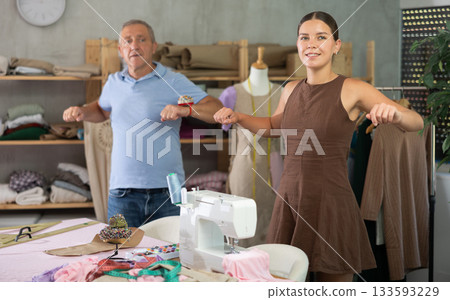 Elderly man and young woman doing warm-up exercises in sewing workshop Elderly man and young woman doing warm-up exercises in sewing workshop 133593229