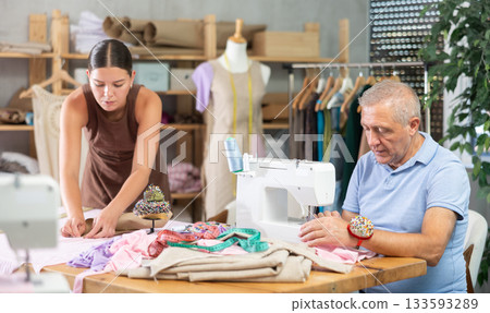 Senior man employee sewing, stitch clothes, girl apprentice draw dress pattern Senior man employee sewing, stitch clothes, girl apprentice draw dress pattern 133593289