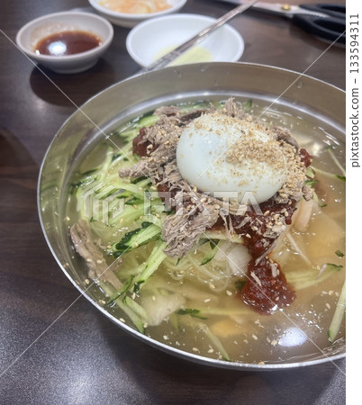 Chilled, chewy naengmyeon noodles in tangy broth paired with juicy, savory mandu dumplings, offering a perfect balance of refreshing coolness and hearty comfort in every delicious Korean bite. 133594311