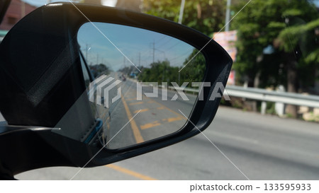Side mirror view of black car. See the yellow lines dividing the lanes of the asphalt road reflected in the mirror. Background of guardrail with trees side of road. 133595933