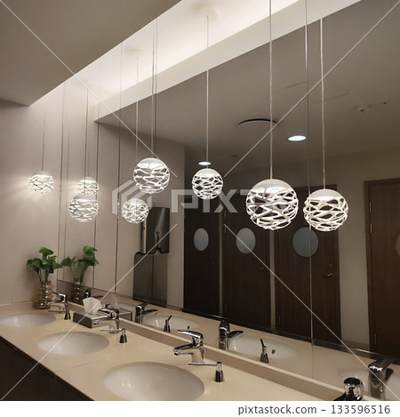 Modern restroom interior with a row of stylish pendant lights featuring intricate lattice designs, reflecting in a large mirror above a beige countertop with integrated sinks 133596516