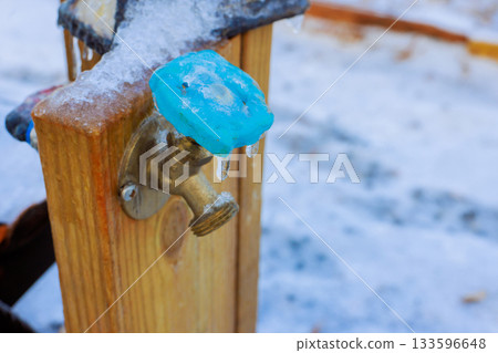 Icy water faucet on wooden post shows signs of freezing in snowy environment during winter. 133596648