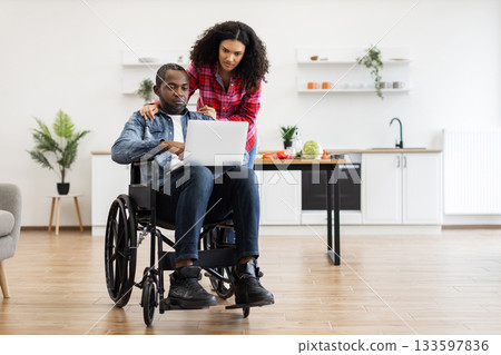 A Black man in a wheelchair and a woman are looking at a laptop in a modern kitchen setting. A Black man in a wheelchair and a woman are looking at a laptop in a modern kitchen setting. 133597836