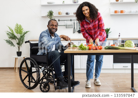 A smiling couple prepares a meal together in a bright, modern kitchen, with one person using a wheelchair. 133597847