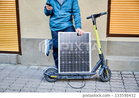 Cropped view of male person holds solar panel, for charging electric scooter, using smartphone. Integration of solar power as sustainable energy source, promoting eco-friendly urban transportation. Cropped view of male person holds solar panel, for charging electric scooter, using smartphone. Integration of solar power as sustainable energy source, promoting eco-friendly urban transportation. 133598432