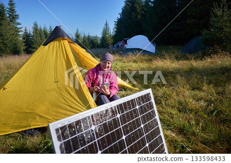 Happy woman sits in front of yellow tent, charging her phone by using solar panel, highlighting use of renewable energy and eco-friendly camping. On background lush green trees and clear blue sky. 133598433