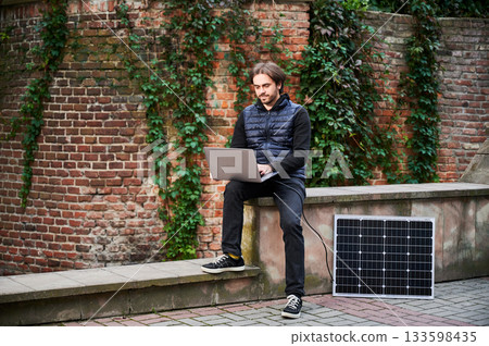 Man sits on stone bench in front of brick wall, working on laptop, connected to solar panel. Urban setting, casual attire highlight modern sustainable, mobile workspace. Man sits on stone bench in front of brick wall, working on laptop, connected to solar panel. Urban setting, casual attire highlight modern sustainable, mobile workspace. 133598435