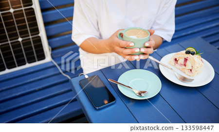 Girl drinking coffee while phone charging from solar panel. Cropped view of woman sitting next to portable solar battery charger. Integration of sustainable renewable energy into everyday life. Girl drinking coffee while phone charging from solar panel. Cropped view of woman sitting next to portable solar battery charger. Integration of sustainable renewable energy into everyday life. 133598447