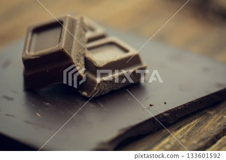 Two dark chocolate squares are resting on slate board over rustic wooden tabletop in flat design Two dark chocolate squares are resting on slate board over rustic wooden tabletop in flat design 133601592