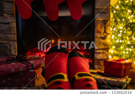 Woman legs with christmas socks and home interior with fireplace and dark wall background. Free space for your decoration. Woman legs with christmas socks and home interior with fireplace and dark wall background. Free space for your decoration. 133602016