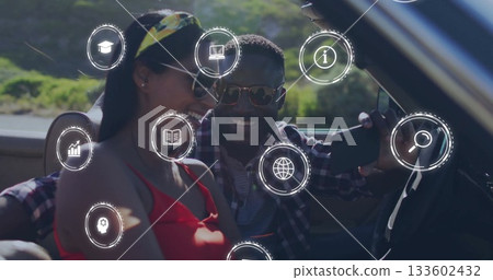 Smiling couple wearing sunglasses using phone in convertible on hillside road, with digital icons 133602432