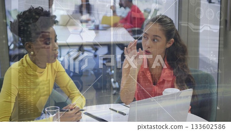 Women in yellow top and pink blouse collaborating at office table, with laptop and digital overlays 133602586