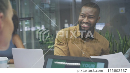 Smiling man in mustard shirt holding document at meeting room table, with laptops and coffee cups 133602587