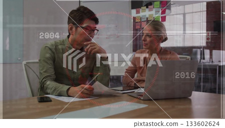 Two colleagues wearing business attire reviewing documents and laptop at office table, with notes 133602624
