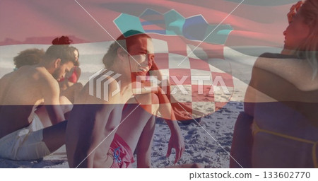 Laughing seven friends wearing swimwear sitting on sandy beach, with turquoise umbrella canopy 133602770