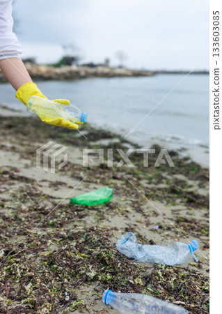 volunteer collecting plastic bottles on a polluted beach, highlighting ocean pollution, waste problem, environmental protection and coastal cleanup action. 133603085