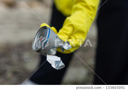 volunteer picking up a crushed metal can during beach cleanup, highlighting pollution, waste problem, ecology, coastal protection and environmental awareness. 133603091