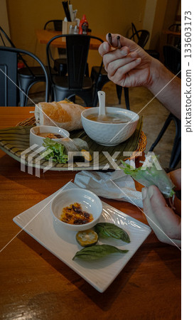 Candid scene with hands preparing fresh rolls or goi cuon and pho bo soup in a cozy Vietnamese restaurant setting Candid scene with hands preparing fresh rolls or goi cuon and pho bo soup in a cozy Vietnamese restaurant setting 133603173