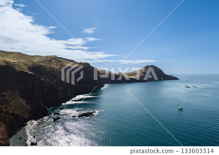 Sao Lourenco cliffs rise over Atlantic as waves trace shore and sailboat floats nearby 133603314