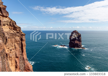 Dark rock stack rises near Madeira cliffs above deep Atlantic waves and glowing sunlight Dark rock stack rises near Madeira cliffs above deep Atlantic waves and glowing sunlight 133603318
