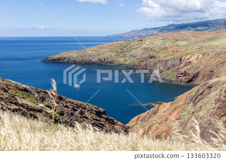 Rugged volcanic shore at Sao Lourenco curves around calm water with sailboat and grass Rugged volcanic shore at Sao Lourenco curves around calm water with sailboat and grass 133603320