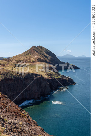 Vertical view of Sao Lourenco cliffs with sunlit slopes above calm clear Atlantic water 133603323