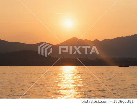 Scenic landscape of West Lake, Hangzhou, during golden hour with sun rays shining through the distant mountains 133603391