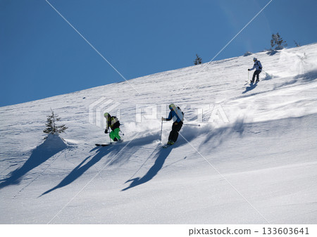 Three skiers carve down a sunny, powder-covered mountain slope, kicking up fresh snow under a clear blue sky, creating a dynamic winter sports action scene Three skiers carve down a sunny, powder-covered mountain slope, kicking up fresh snow under a clear blue sky, creating a dynamic winter sports action scene 133603641