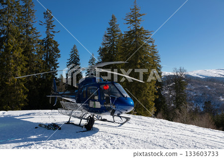 A blue helicopter awaits on a snowy mountain, ready for heli-skiing. Surrounded by pine trees and stunning alpine scenery under a clear blue sky, it captures the essence of winter adventure 133603733