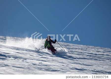 A female skier carves through fresh powder on a sunny mountain slope, kicking up snow as she descends, creating a dynamic and energetic winter sports action scene under a clear blue sky A female skier carves through fresh powder on a sunny mountain slope, kicking up snow as she descends, creating a dynamic and energetic winter sports action scene under a clear blue sky 133603734
