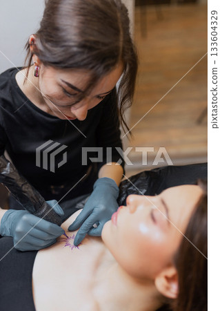 tattoo artist working on a woman chest design in a studio, showing the process of professional tattooing, precision, sterile gloves and cosmetic procedure atmosphere. tattoo artist working on a woman chest design in a studio, showing the process of professional tattooing, precision, sterile gloves and cosmetic procedure atmosphere. 133604329