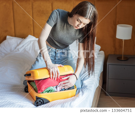 Young woman struggling with overstuffed suitcase on bed 133604751