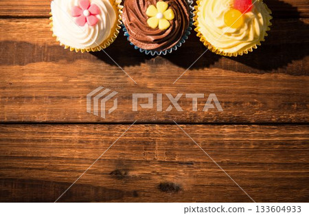 Three frosted cupcakes are resting on wood grain table in flat design with fondant flowers, gummies Three frosted cupcakes are resting on wood grain table in flat design with fondant flowers, gummies 133604933