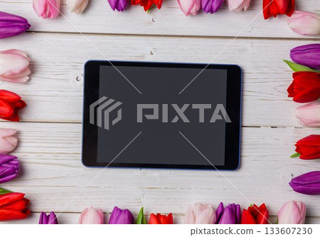 Rectangular tablet is being shown in flat lay on painted wooden tabletop, with tulip floral border 133607230