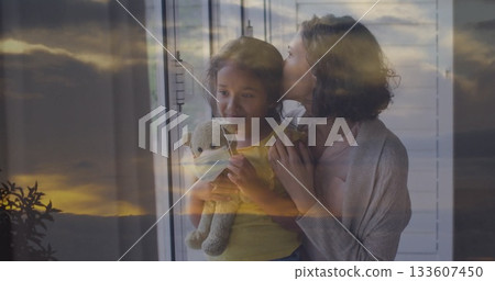 Mother embracing daughter wearing yellow top at home window, with gray teddy bear mask Mother embracing daughter wearing yellow top at home window, with gray teddy bear mask 133607450