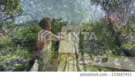 Adult artist wearing apron painting blank canvas in lush garden, with paint jars and brushes 133607451