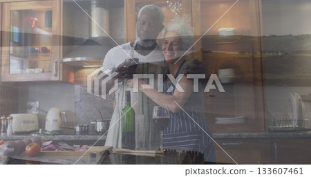 Toasting couple wearing aprons cooking in home kitchen, with wine glasses, cutting board, knife Toasting couple wearing aprons cooking in home kitchen, with wine glasses, cutting board, knife 133607461