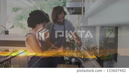 Cooking couple in aprons chopping vegetables and stirring pot at kitchen counter with sunset glow 133607482