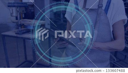 Holding tablet tech in grey apron using touchscreen at workbench, with drill press and shelves 133607488