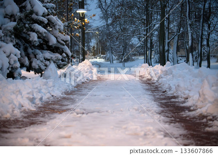 Snowy Park Path at Dusk with Lamplight and Snow Piles Along the Walkway Snowy Park Path at Dusk with Lamplight and Snow Piles Along the Walkway 133607866