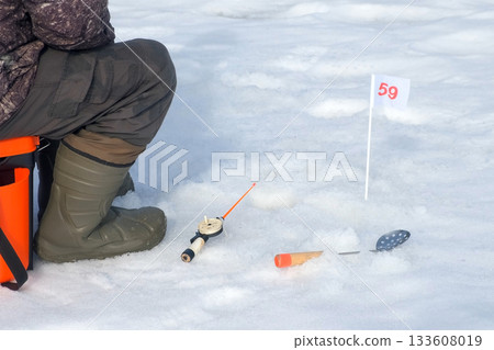 Ice fisherman sitting patiently with rod, flag indicator on fishing competition. Ice fisherman sitting patiently with rod, flag indicator on fishing competition. 133608019