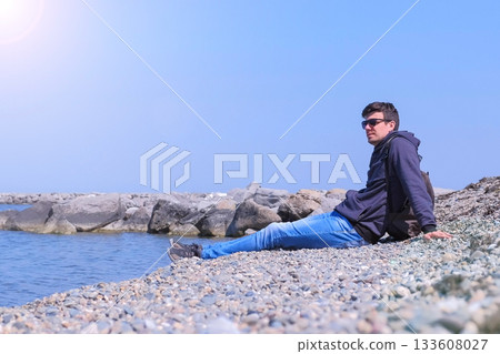 Man tourist traveller comes to stone beach sits and looks at sea. Man tourist traveller comes to stone beach sits and looks at sea. 133608027