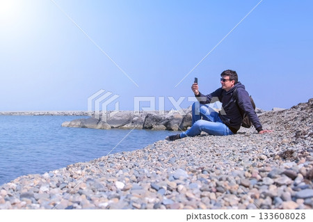 Guy talks video chat in his smartphone sits on sea stone beach on vacation. Guy talks video chat in his smartphone sits on sea stone beach on vacation. 133608028