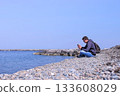 Man traveller is sitting on sea stone beach and browsing smartphone, side view. 133608029
