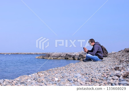 Man traveller is sitting on sea stone beach and browsing smartphone, side view. Man traveller is sitting on sea stone beach and browsing smartphone, side view. 133608029