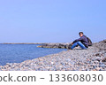 Young man traveler sits on stone beach and looks at sea on spring vacation. 133608030